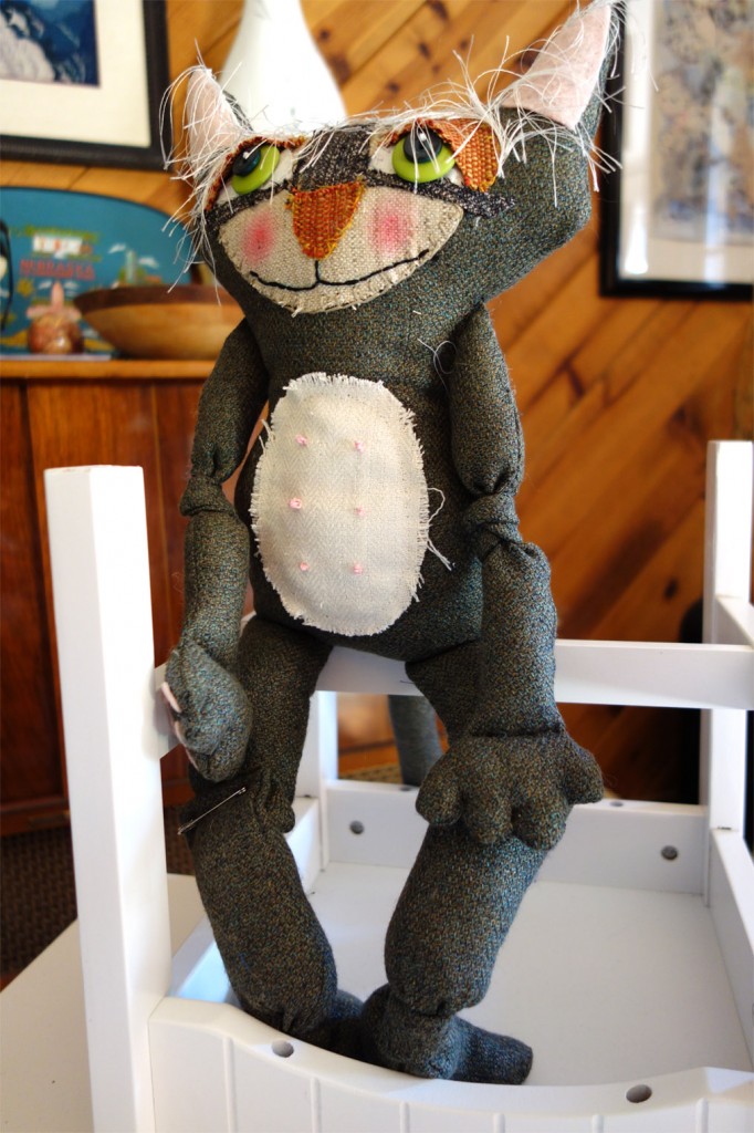 Old whimsical cat doll sits on upside down chair on table top.
