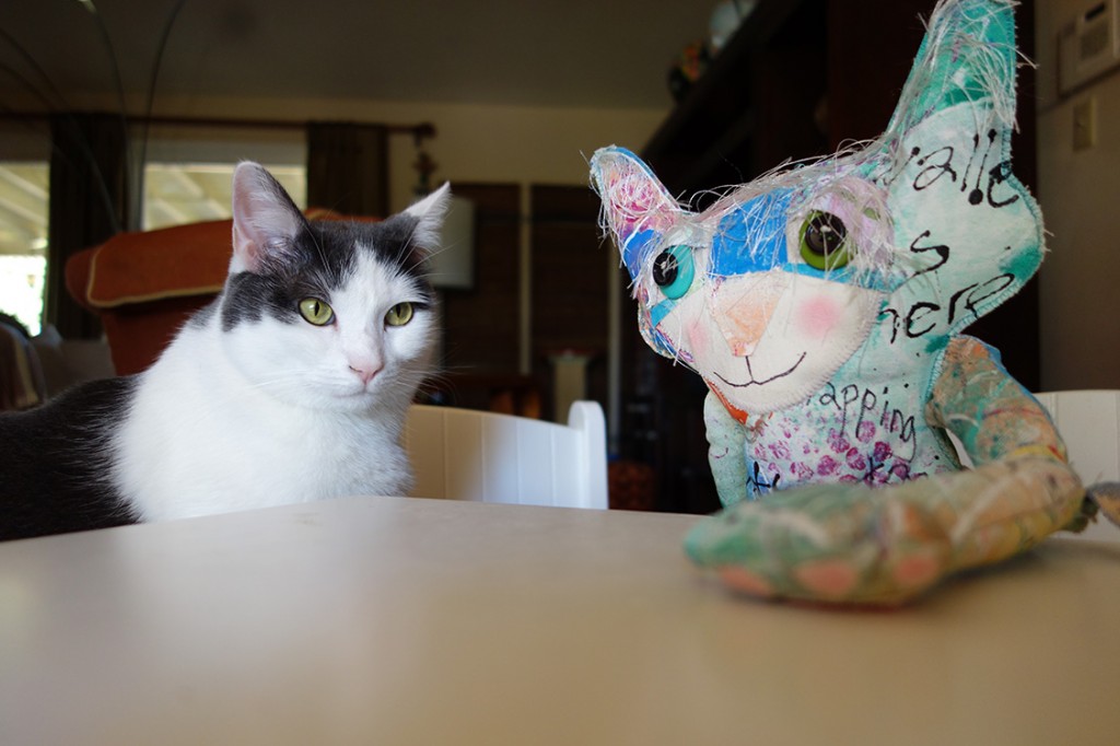 Wally the real cat and a whimsical cat doll sit at table.