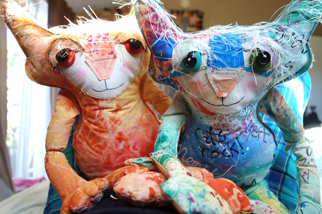 Two adorable soft-sculpture cat dolls sit on the bed staring on their human.