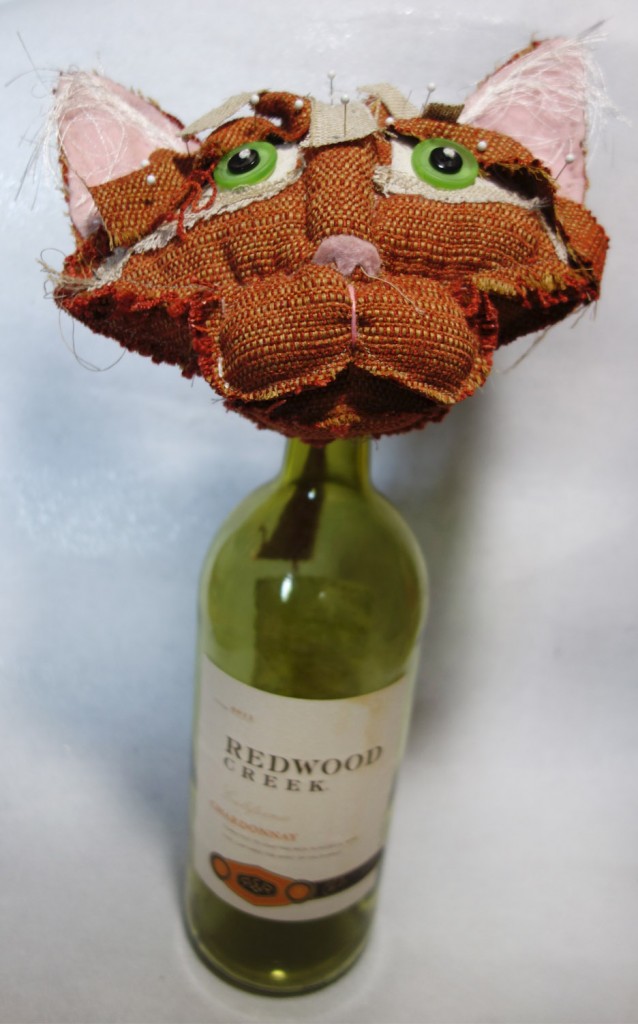 Orange soft-sculptured cat doll head propped on top of an empty large wine bottle. Pins are holding fabric in place.