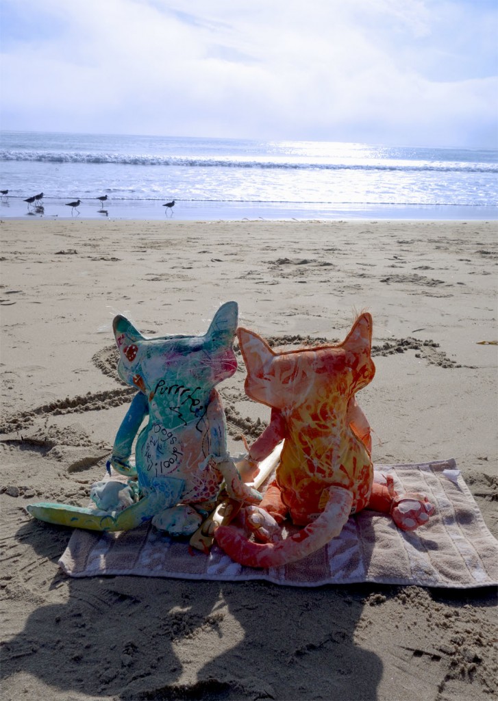 The Chairman and Ginger, two cute cat dolls stare out at the ocean as a flock of sandpipers runs through the wet sand.