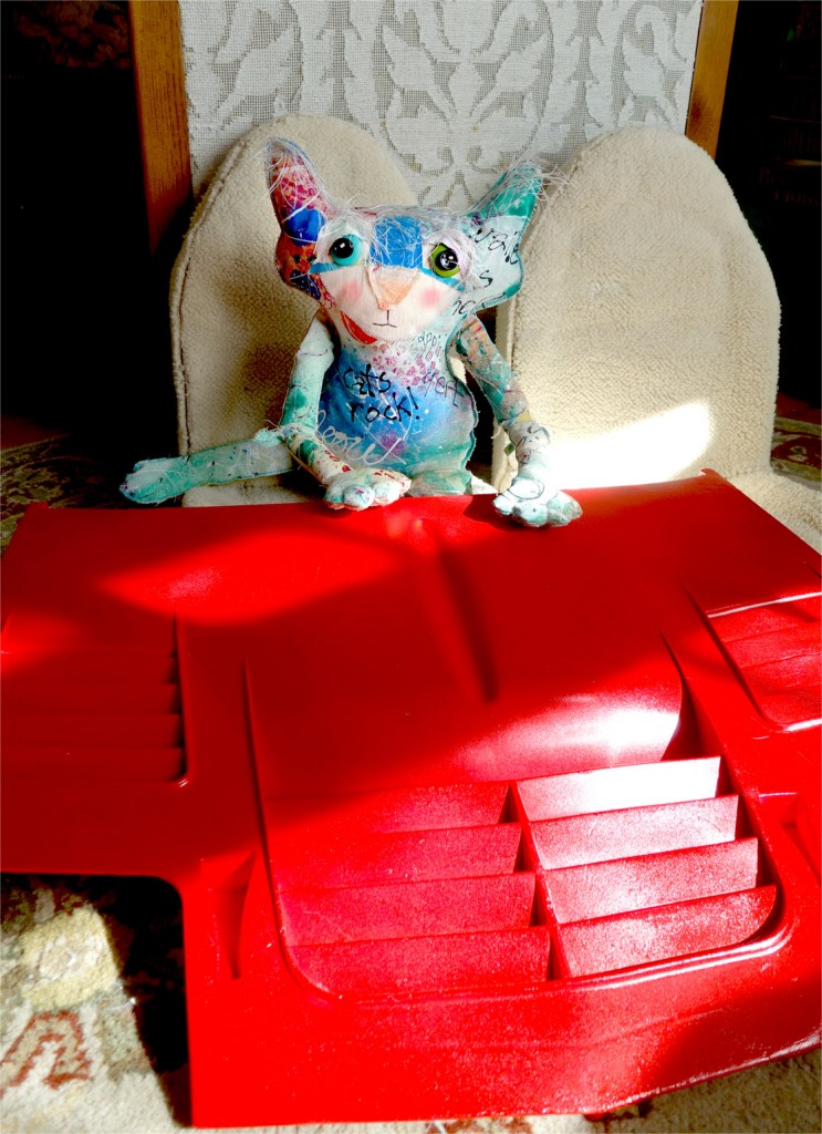 The Chairman cat doll is sitting on a seat taken out of his car, with a bright red car hood on the floor in front of him. The hood looks great!