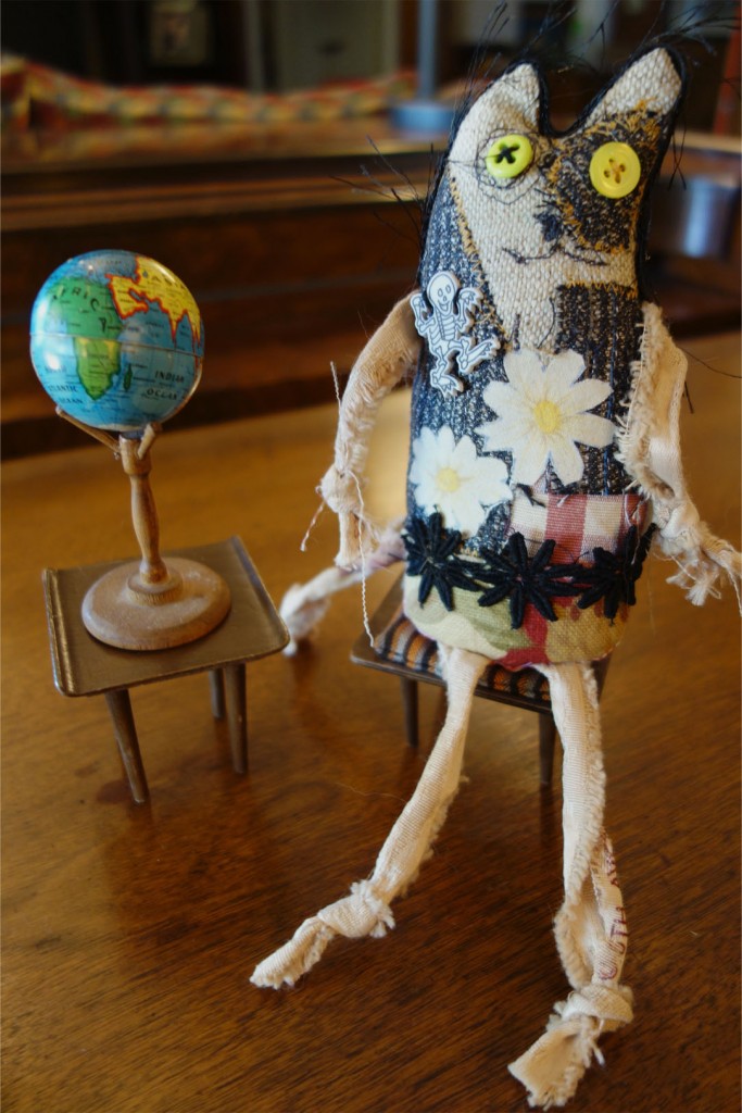 Freaky little cat doll sits on a mid-century modern piece of doll furiture with a small, metal globe of earth sitting next to him.