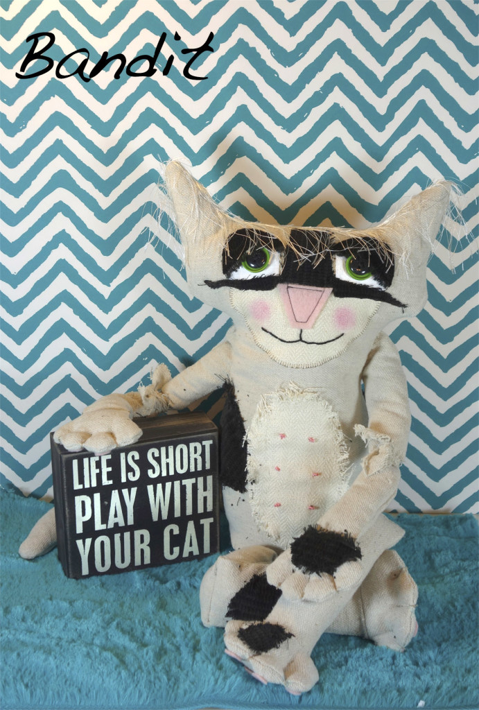 Adorable soft sculpture cat doll with large black velvet spots. He sits on a turquoise rug with turquoise and white wallpaper in the background. He poses with a sign that reads: life is short. Play with your cat.