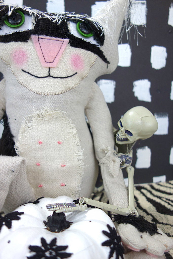 Adorable soft sculptured cat doll named Bandit. He poses with a black and white pumpkin. A skeleton is sneaking around from behind him to grab the pumpkin.