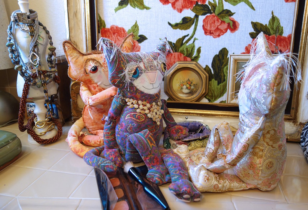 Three adorable soft sculpture cat dolls and are a bathroom counter with makeup and jewelry. Ginger is fastening a necklace around Eve's neck. Peach looks on.