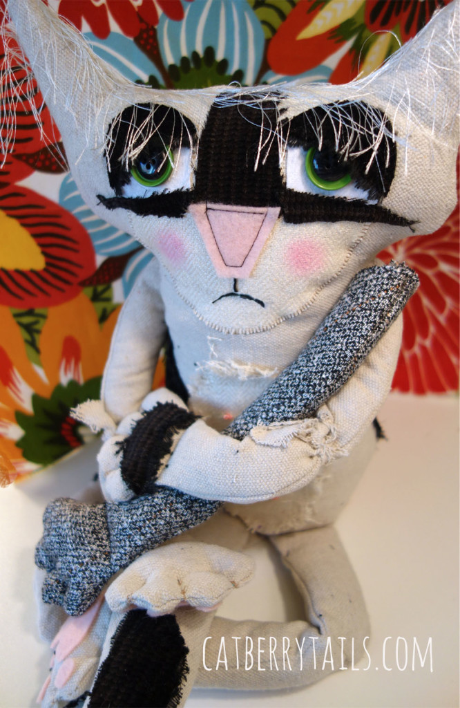 Bandit, the very cute soft sculptured cat doll, has a frown on his face as he is grasping a cat doll arm.