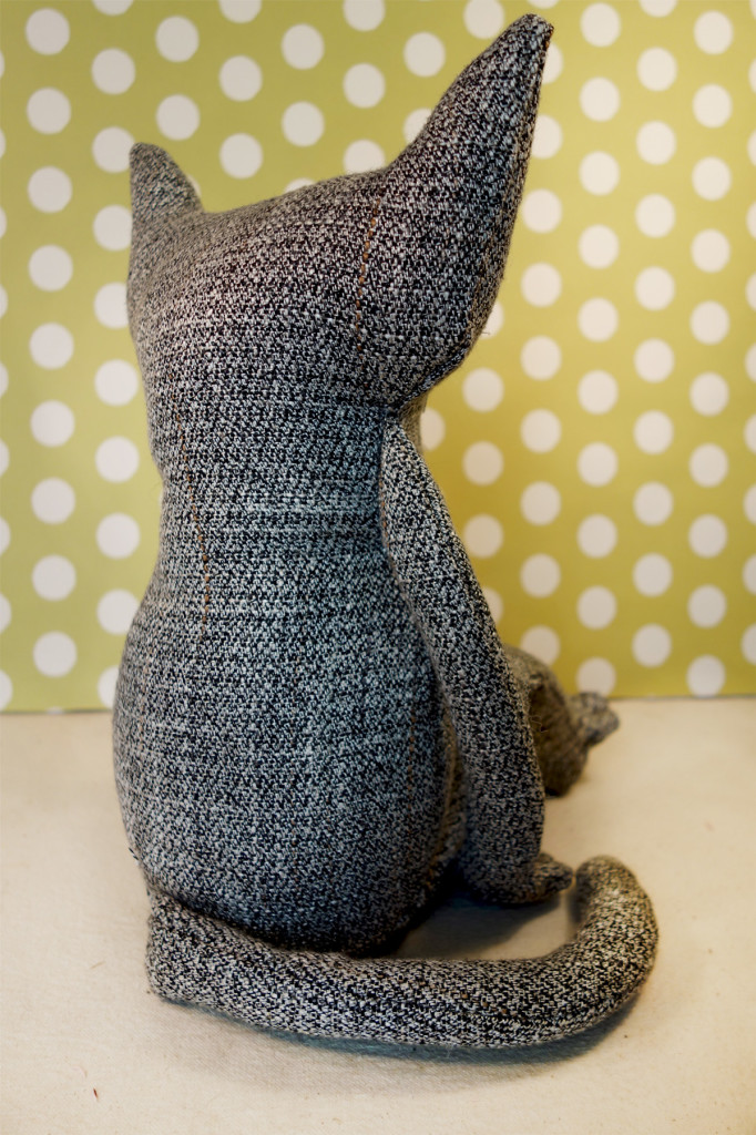 A brand new soft sculpture cat is featured in a photograph. He is seated with with his back to us and his tail wrapped around. He's made out of a man's charcoal grey tweed suit.