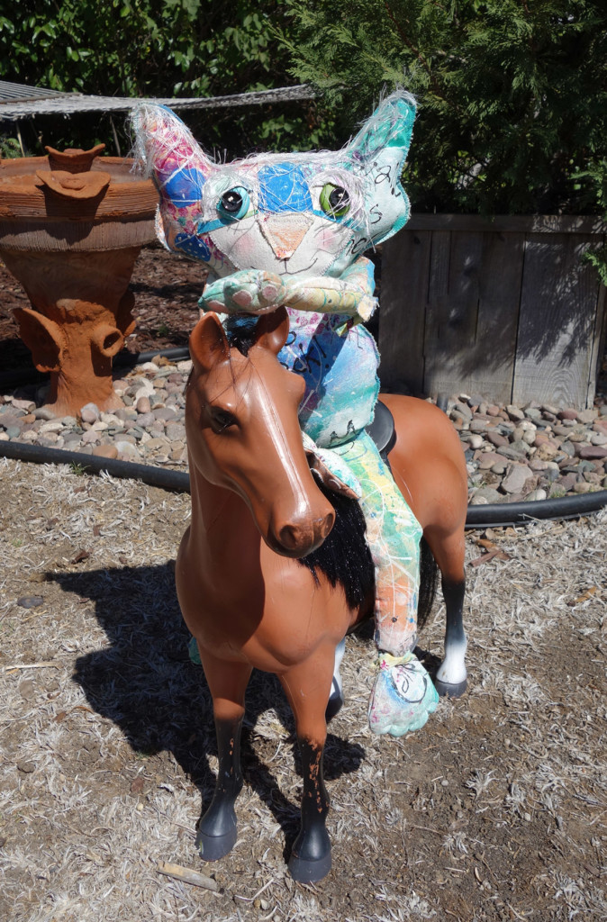 The Chairman, whimsical cat doll, sits on a large plastic toy horse in a sunny back yard.